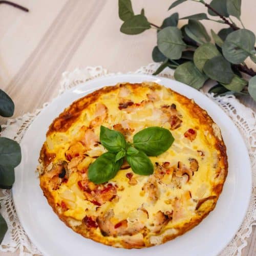 kana-ananas-piirakka (4) Koko piirakka pitsireunaisella lautasella, taustalla vihreitä oksia ja kodikas tunnelma.