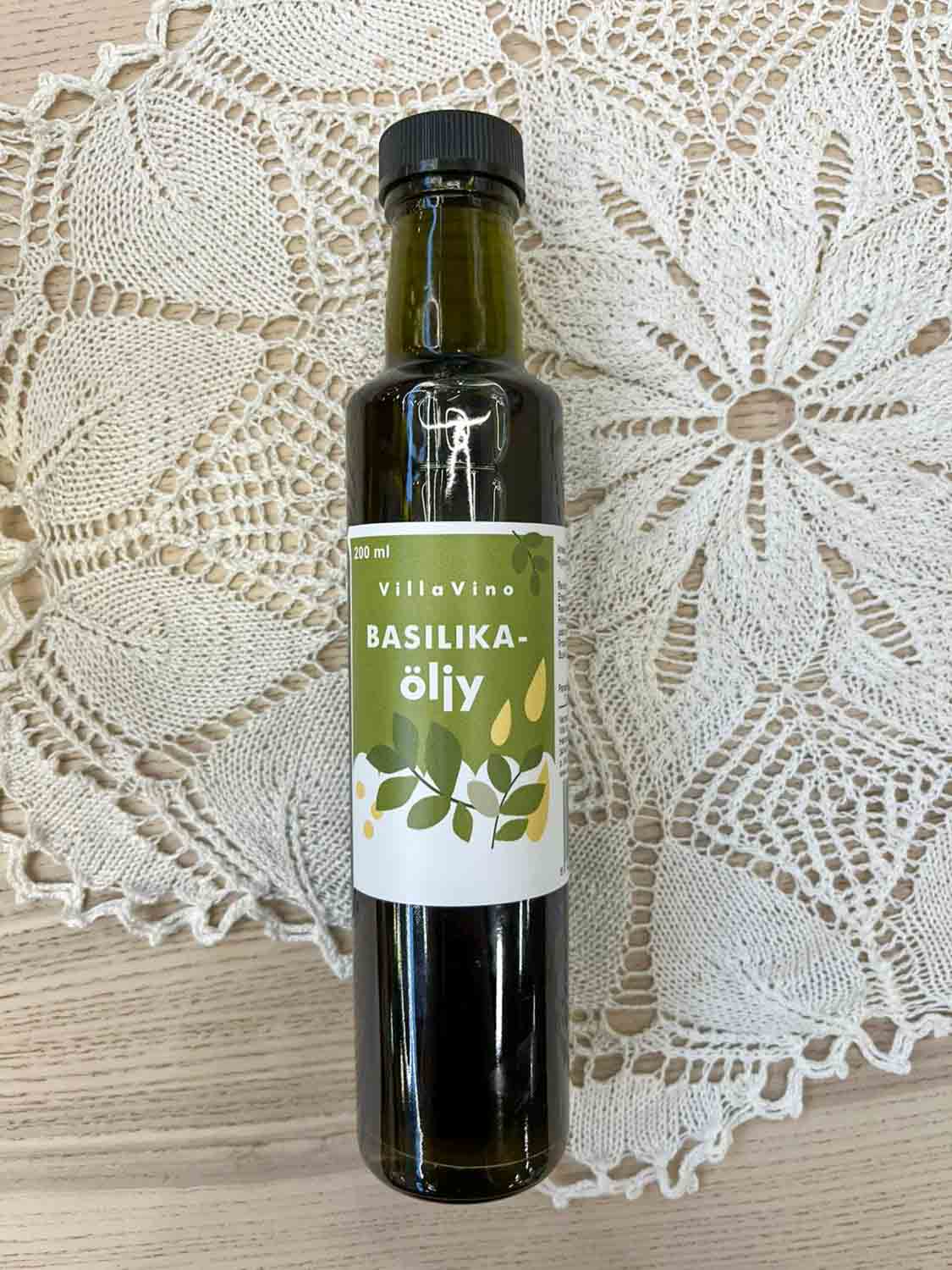 Villa Vinon Basilikaöljy kauniissa lasipullossa jossa vihertävä etiketti. Pullo on puupöydällä pitsiliinan päällä.