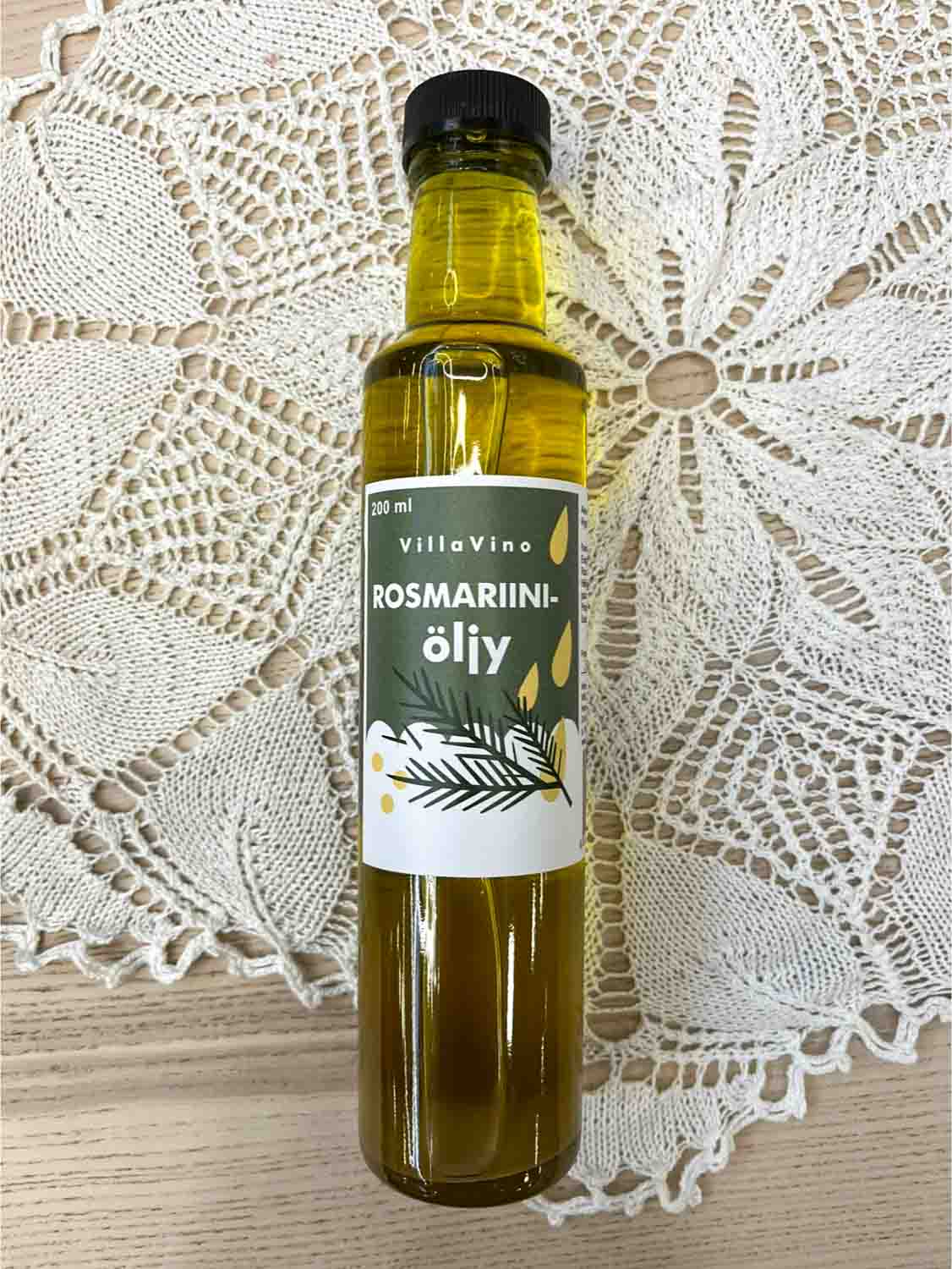Villa Vinon Rosmariiniöljy kauniissa lasipullossa jossa vihertävä etiketti. Pullo on puupöydällä pitsiliinan päällä.