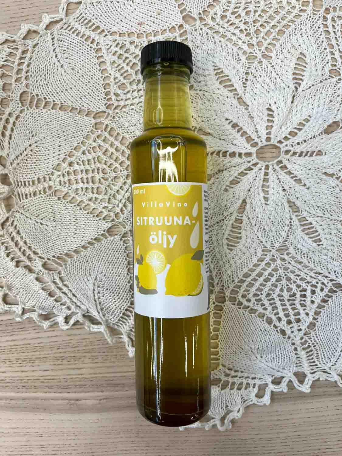 Villa Vinon Sitruunaöljy kauniissa lasipullossa jossa keltainen etiketti. Pullo on puupöydällä pitsiliinan päällä.