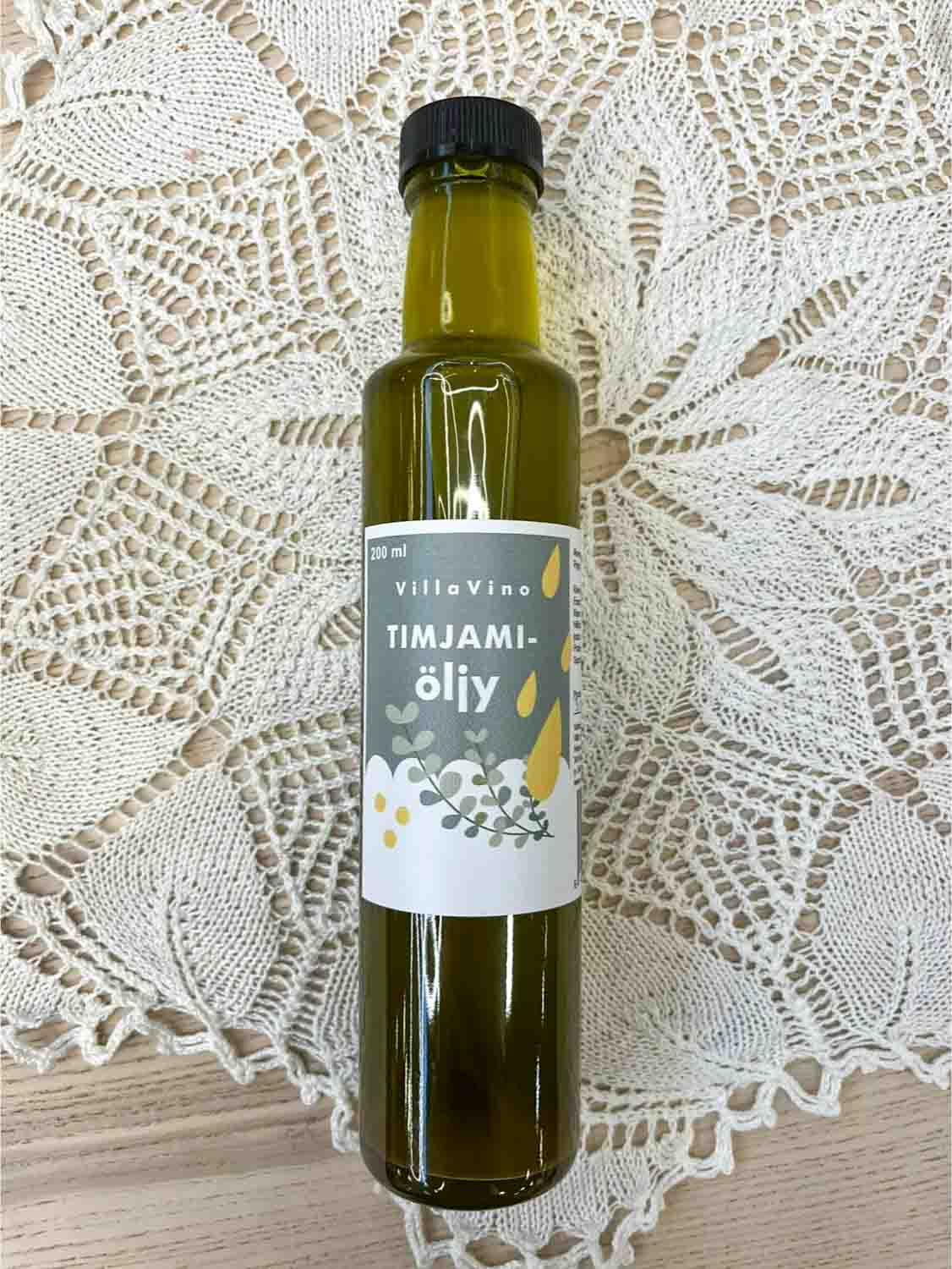 Villa Vinon Timjamiöljy kauniissa lasipullossa jossa vihertävä etiketti. Pullo on puupöydällä pitsiliinan päällä.