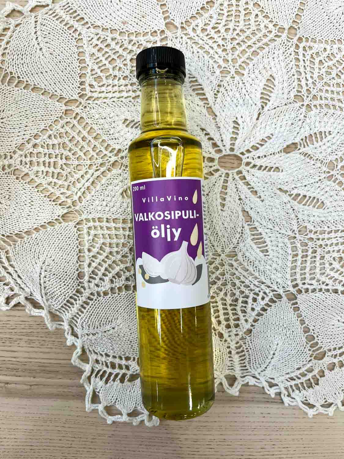 Villa Vinon Valkosipuliöljy kauniissa lasipullossa jossa violetin sävyinen etiketti. Pullo on puupöydällä pitsiliinan päällä.