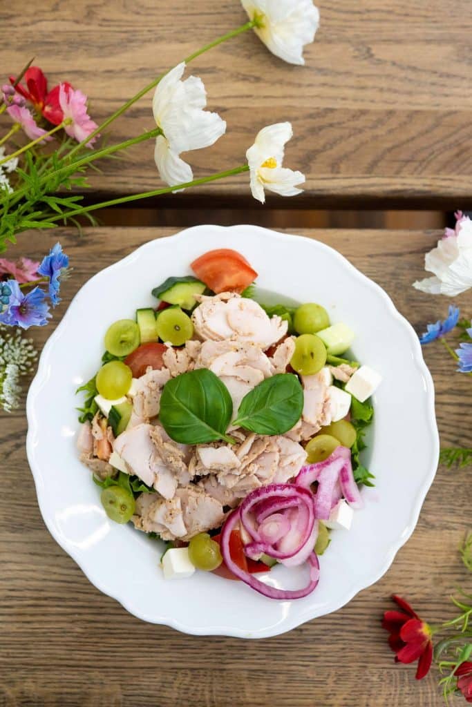 Villa Vinon kesäkaffilan maukas ja värikäs salaattiannos valkoisella lautasella, Puinen pöytätaso ja niittykukkia.