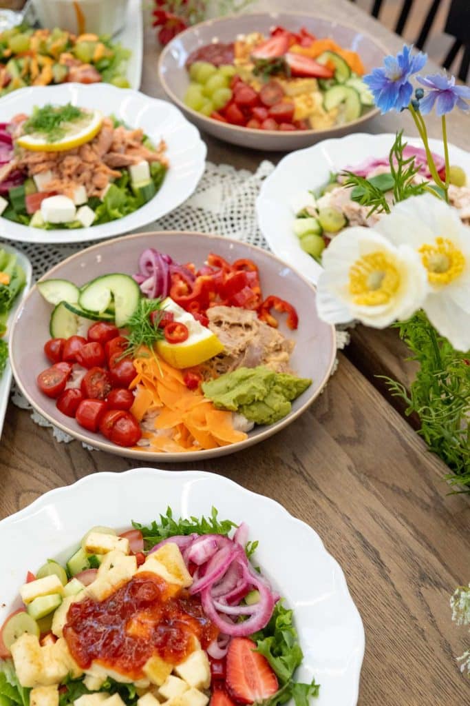 Värikkäitä Villa Vinon kesäkaffilan bowl- ja alaattiannoksia valkoisilla ja vaaleilla lautasilla. Alla puinen pöytätaso ja koristeena niittykukkia