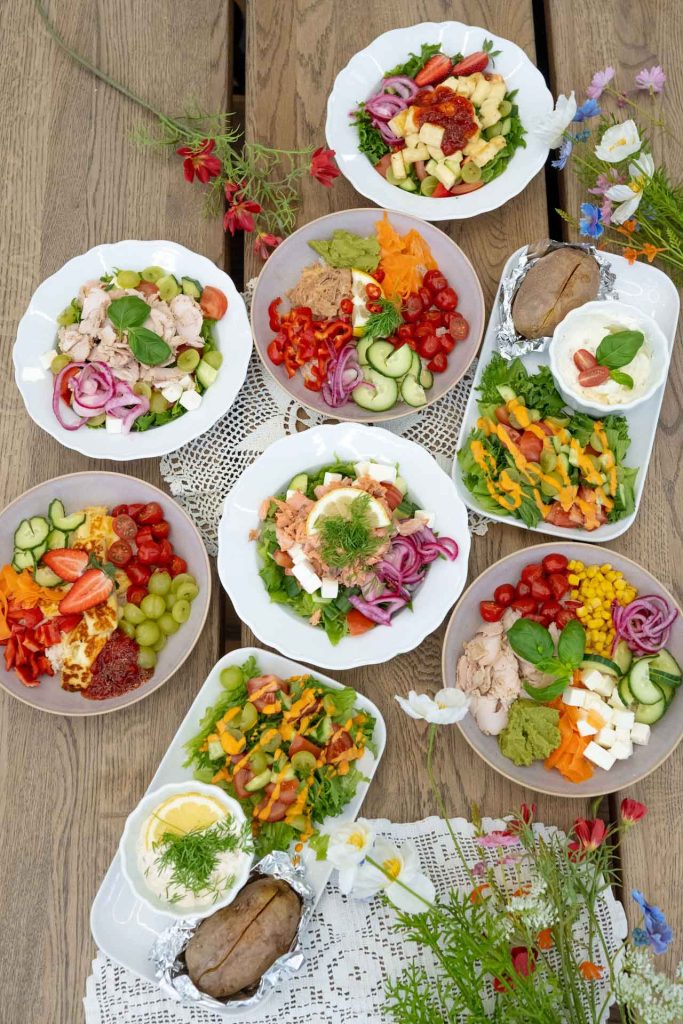 Värikkäitä Villa Vinon kesäkaffilan bowl- ja alaattiannoksia valkoisilla ja vaaleilla lautasilla. Alla puinen pöytätaso ja koristeena niittykukkia